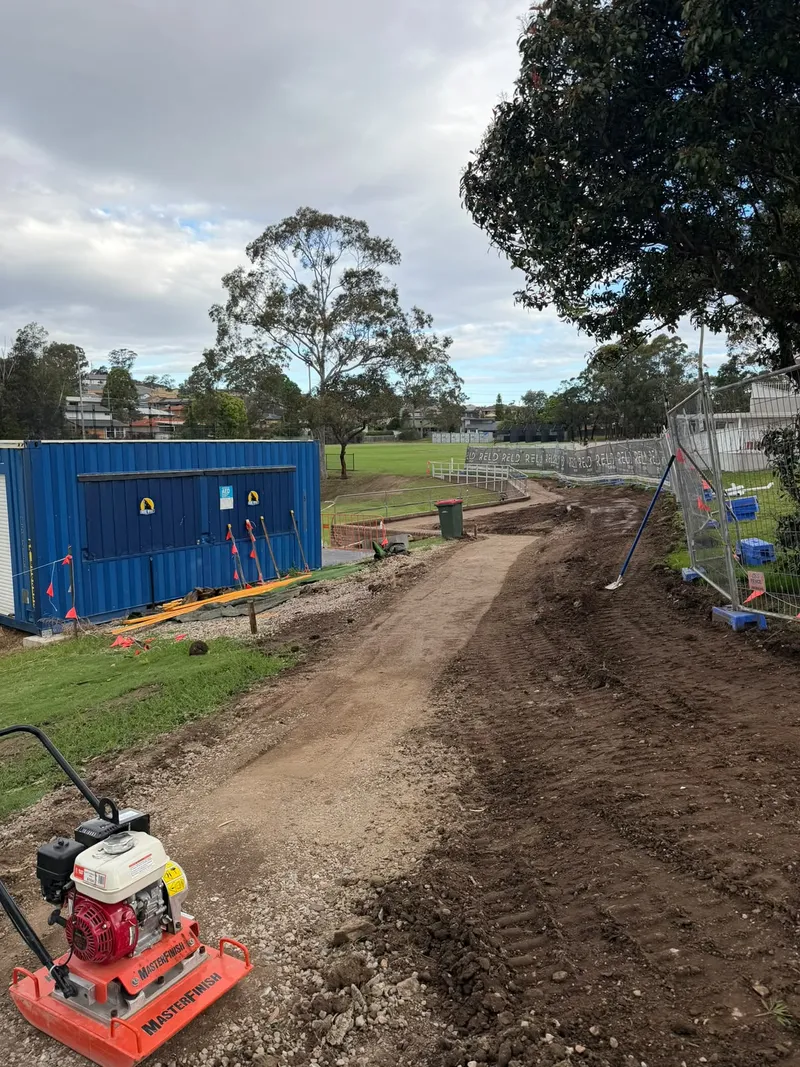 The excavator bucket on site at Max Ruddock Reserve, with the blue site office and oval behind
