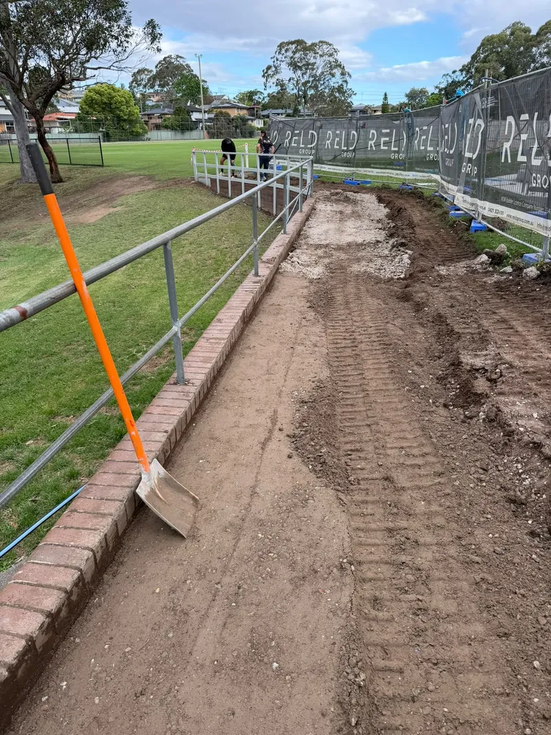 A shovel and the excavated trench along the guard rail during site preparation, with the crew working behind the fencing