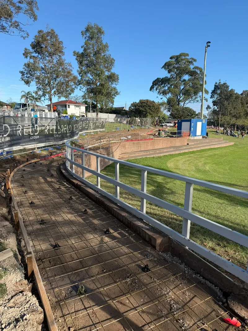 Steel mesh laid in formwork along the full curve of the path beside the oval, with the guard rail and brick edging in place